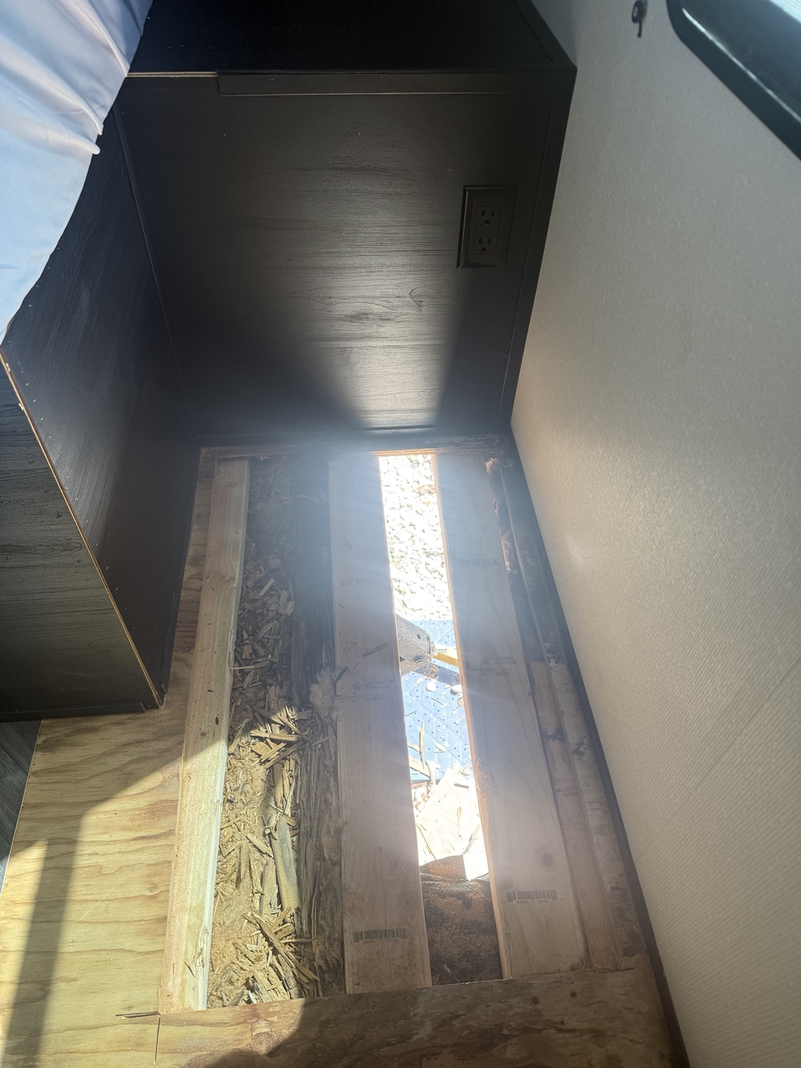 Opened floor beside the RV bed, showing the exposed subfloor area.